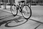 A black-and-white photograph of a cyclist riding through an urban plaza, captured with motion blur. Strong sunlight casts a large, sharply defined shadow of the bicycle and rider across the paved ground.