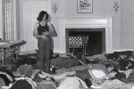 A black-and-white archival photograph shows a woman standing in the center of a room while a group of people lie on the floor around her, suggesting a guided workshop or consciousness-raising session.