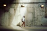 Person wearing headphones and a backpack walks along a sunlit concrete wall in an urban street. Person wearing headphones and a backpack walks along a sunlit concrete wall in an urban street.