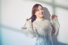 A woman in a white lace dress with long brown hair smiles and poses against a white backdrop.
