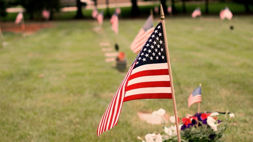 A small American flag staked in the ground with green grass and the corner of a gravestone in the background.