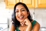 Smiling woman with shoulder-length dark hair leaning on a kitchen counter, wearing a green sleeveless top and a gold necklace, with light wood cabinets in the background. Smiling woman with shoulder-length dark hair leaning on a kitchen counter, wearing a green sleeveless top and a gold necklace, with light wood cabinets in the background.