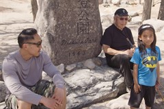 Two men sit on either side of a stone inscribed with Japanese characters and "Pleasure Park 1943." A child stands in front.