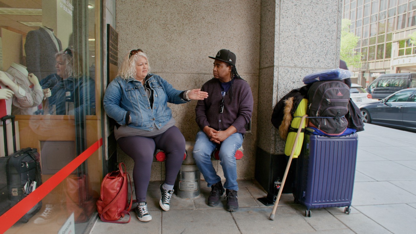 Two seated people face each other on a sidewalk near a building. One gestures towards the road. There is a suitcase and other items stacked nearby.