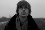 Close-up of a wide-eyed young white man wearing a coat and scarf before a stark background