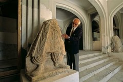 Man speaking next to a large broken statue, of which only the legs and feet of a figure remain. Man speaking next to a large broken statue, of which only the legs and feet of a figure remain.