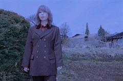 Blue-toned photo of a woman in a suit, in front of a building surrounded by a barbed-wire fence.