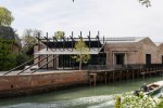 A waterfront complex of historic brick buildings is joined by a contemporary black steel pavilion and covered terrace. A canal runs alongside the buildings, where a small motorboat passes in the foreground.