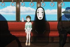 A girl sits in a train car next to a see-through spirit in a white mask. The windows look out on water and blue sky.