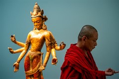 A man in red robes with shorn hair and a crowned three-armed sculpture faces opposite ways, hands posed for meditation.