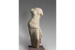 A fragmentary sculpture in white marble depicts a woman’s body draped in fabric below the waist, with significant breaks around the neck, shoulders, and knees.