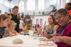 Adults and a child sit around a table in a room with high ceiling and stained-glass windows with various art tools in front of them.