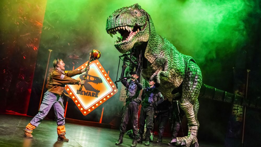 A performer stands in front of a life-like T-Rex that is controlled by two other performers