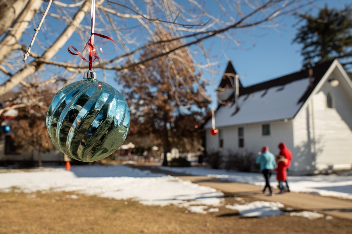 blue shiny ornament hangs from a tree with two people walking past an old church in the background