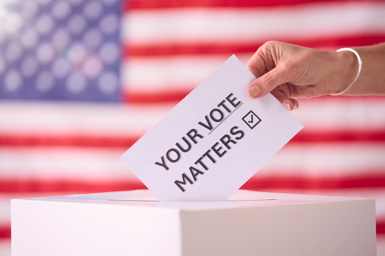 Hand placing a card into a box. The card says, your vote matters. An American flag is in the background.