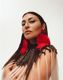 Portrait of musician Elisapie, her dark brown hair blends into a tied and fringed top and she wears large red fringed earrings.