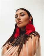 Portrait of musician Elisapie, her dark brown hair blends into a tied and fringed top and she wears large red fringed earrings. Portrait of musician Elisapie, her dark brown hair blends into a tied and fringed top and she wears large red fringed earrings.