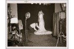 Photograph of a woman in a bridal gown in a photography studio, with a man kneeling on the ground arranging the train of the dress.