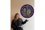 In this photomontage, a young woman gestures toward a round stained glass window that appears to float. It has a blue background and shows two small groups of men gathered in front of a narrow column.