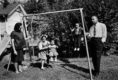 Black and white photograph of Dr. Martin Luther King, Jr and Coretta Scott King standing on either side of swing set. Three of their children are playing on the swings.