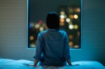 person alone in bedroom looking out of a window at a city