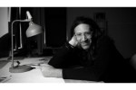 A black and white photograph of a man wearing glasses and a black shirt seated at a desk with a lamp. He is resting his cheek against his hand and looking at the camera.