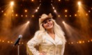 A female singer with blonde hair visible from waist up holding a microphone and wearing a white and golden country hat and outfit. In the background is multiple golden spotlights. A female singer with blonde hair visible from waist up holding a microphone and wearing a white and golden country hat and outfit. In the background is multiple golden spotlights.