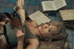 a blond white woman in a tank top lies on the floor with open books scattered around, reading one