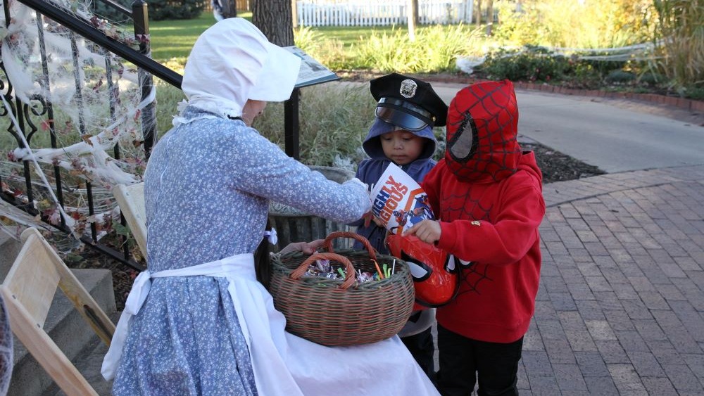 Child in spiderman costume and child in police officer costume reach for candy in a basket held by woman in pioneer costume