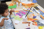 A child holding two paint brushes applies glue to an artwork, already covered with scraps of colorful tissue paper and black and white photographs, on the table in front of him.