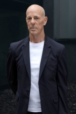 Serious-looking bald man in a dark blazer and white shirt standing against a plain, dark background.