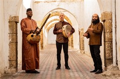 Three men stand in a hall of archways, each holding an instrument: a kora, a handheld drum, and a setar.